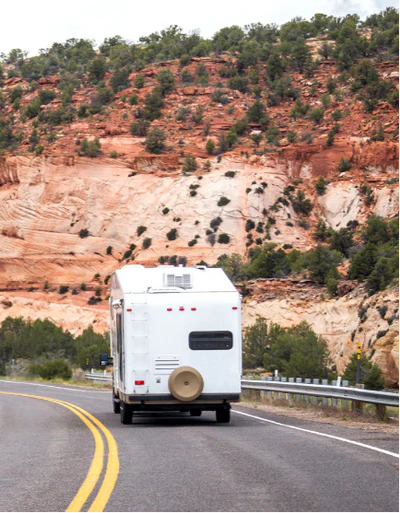 White RV on a road with red rock cliffs and greenery in the background